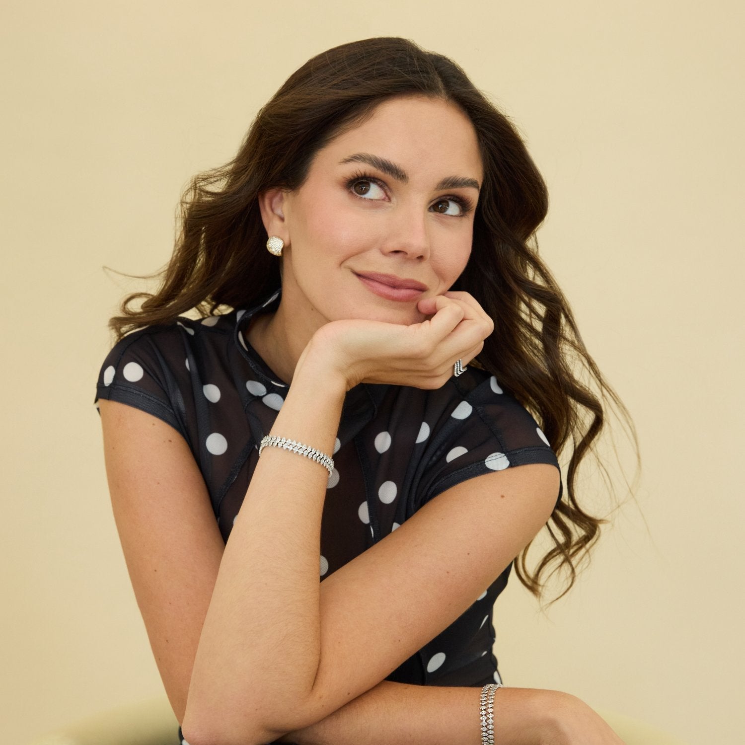 Woman wearing a black dress with white polka dots, sitting on a chair against a beige background.